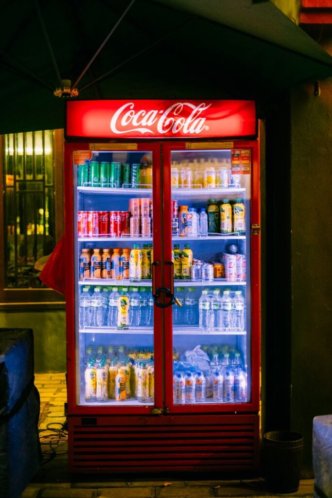 Stocked Coca-Cola fridge filled with assorted beverages on a night street corner.